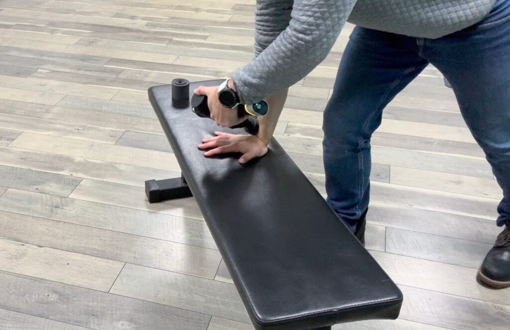 Dr. John with his wrist placed on a bench in a modified push up position using a massage gun into the crease of the wrist